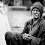 A black and white portrait of an elderly man sitting on the streets, expressing hardship and resilience.